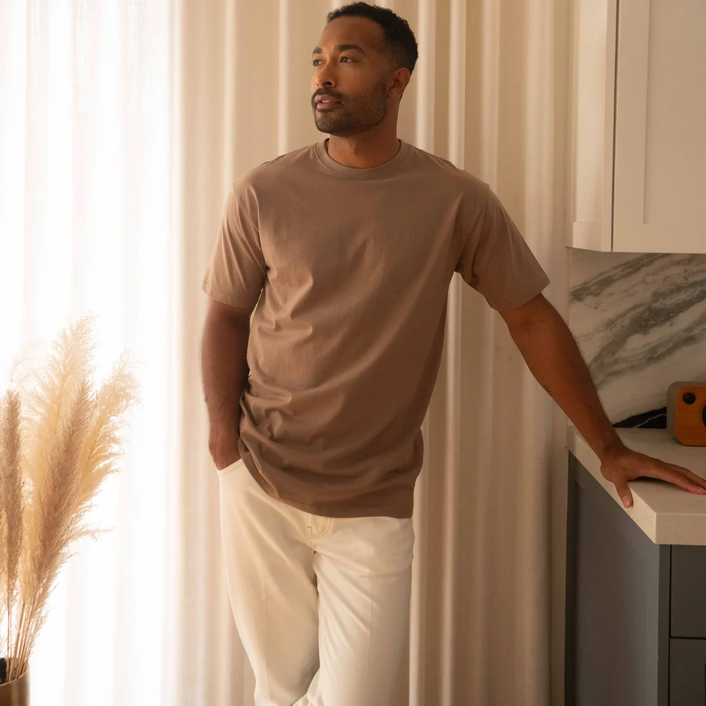 Man wearing a latte Cotton Heritage MC1086 men's heavyweight T-shirt with cream pants in a modern interior.